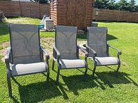 Front view of three identical wide patio chairs on grass with a wooden shed in background. Shows mesh seats and metal armrests.