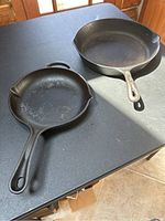 Two cast iron skillets on black table, about 14 inch and 10.5 inch in size, showing handles and pouring spouts.