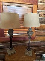 Two oil rubbed bronze decorative lamps with burlap shades sitting on a table in front of a window with wood blinds. Lamps have scroll design and square bases.