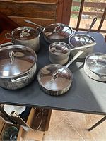 Top-down view of all seven pieces spread out on a black tabletop showing variety of pot and pans with lids