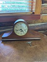 Front view of wooden mantel clock with white face, black numbers and hands, placed on brown surface. Winding key positioned beside clock.