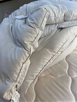 Photo showing close up of off white comforter with visible creasing and tags, folded on a mattress.