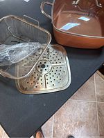 Square copper-colored cooking pot with two handles, a metal frying basket wrapped in plastic, and a perforated metal insert on a dark surface.