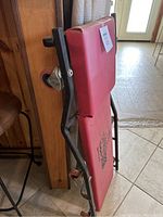 Red padded mechanics creeper leaning against a wooden surface, showing side view with wheels and handle.