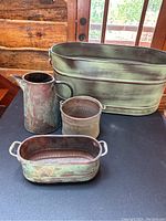 Full view of all four copper pieces on a black table, showing the large oval tub, pitcher, oval container, and round container with patina finish.