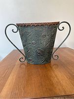 Front view of green patina metal basket showing embossed leaf pattern, woven rim and scroll handles on wood surface.