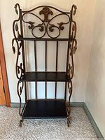 Front view of three-shelf unit showing ornate metal scroll sides and wood shelves, placed in corner indoors on carpet.