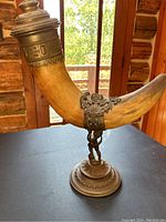 Full view of decorative urn showing curved painted wood body with antique bronze base and lid, set on table with wood-paneled room background.