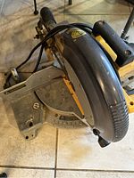 Side angle of the DeWalt mitre saw showing blade cover, power cord, and mitre angle gauge on a tiled floor.