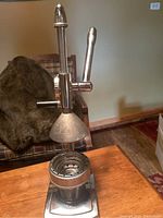 Photo showing vintage metal juice press with lever and base on wooden table, background includes furniture and carpet