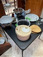 Photo of multiple kitchen gadgets on a black table including a cream-colored salad spinner, metal colander, white ceramic egg dish, gravy boat, measuring cup, watermelon slicer, woven trivets, plastic pitcher, and spoon holder.