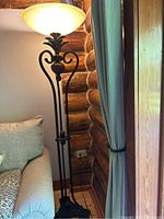 Full view of large standing lamp showing tall metal base with scroll decorative elements and off-white glass shade illuminated.