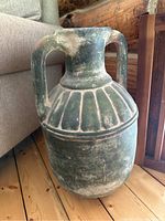 Front-facing photo of a large greenish pottery jug with two handles, standing on a wooden floor next to furniture. The jug has vertical grooves and shows signs of wear with dust and discoloration.