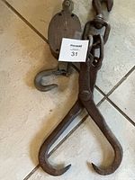Photo of an antique log hook with two sharp prongs alongside a metal block and tackle with pulley and chain links, all showing signs of rust and heavy use.