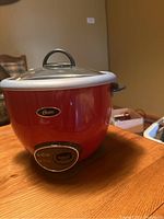 Front view of red Oster electric rice cooker with control panel and glass lid, placed on wooden surface.