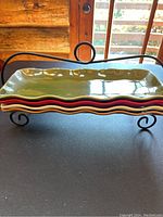 Photo of four stacked glass trays in red, white, yellow, and green colors set in an oil rubbed bronze metal holder with decorative scroll legs and round handle.