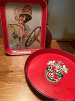 Photo showing rectangular Coca-Cola metal serving tray with red border and vintage girl driving a car image, and round Beck's metal serving tray with Beck's crest logo and 'Product of Germany' text.