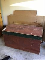 Front angled view of red-painted wooden trunk with dark green band showing wear and scratches.