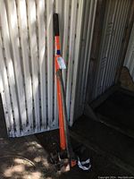 Full view of the invasive plant puller and axe leaning against a corrugated metal wall.