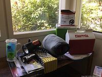 Photo showing two coolers stacked (green Coleman Party Stacker cooler and red Igloo Playmate cooler), tent light box, sleeping bag, flashlights, and grill parts on a table by window.
