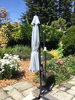 Photo of the full closed umbrella and stand in a garden setting showing light gray fabric and curved black metal stand.