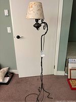 Full view of the black wrought iron floor lamp standing against a white door and sage-green wall, showing the tall stand with curved detail and fabric lampshade.