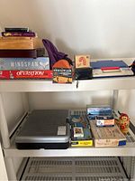Photo showing boxed board games Wingspan, Operation, a candy cane box, orange and black box, and blue and white electronic toy with handle, plus wooden figure and nest doll on shelves