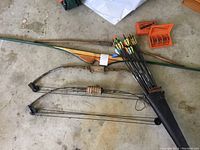 Full view of two bows (64" and 40"), traditional wooden longbow, quiver with arrows, and a box of stainless steel broadheads on concrete floor.
