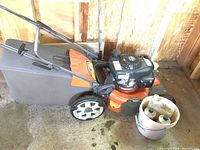 Front-right angled view of mower showing orange deck, gray grass catcher, and a bucket with oil containers beside it