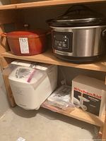 Lot shelf with four kitchen appliances including the Hamilton Beach crockpot, Wolfgang Puck Dutch oven, Black & Decker bread maker, and boxed Kitchen Aid food grinder