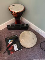 Photo showing African style djembe drum, hand drum, Tin Whistle book, black flute and whistles on carpeted floor.