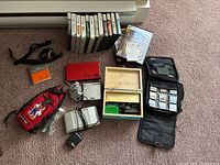 Red Nintendo DS Lite console, charger, straps, pouches, wooden box, and zip case with cartridges arranged on carpet