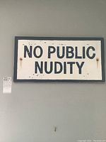 Full frontal view of rectangular wooden sign with bold black text 'NO PUBLIC NUDITY' on white background with black border.