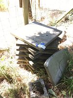 Stack of approx 10 square concrete patio pavers outdoors by a wire fence in grass.