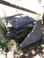 Stack of square concrete patio pavers with one triangular corner piece leaning against the stack outdoors on grass under shade.