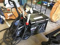 Simoniz pressure washer unit with attached hose and original box in background, placed on a garage floor with other items around.