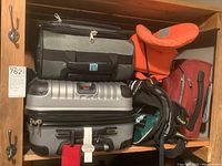 Inside a wooden cabinet showing black Samsonite soft cooler bag, red Delsey suitcase, black Adidas backpack, orange Helly Hansen child life jacket, and silver Wenger suitcase.