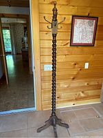 Full view of the vintage wooden coat rack showing the twisted pole, hooks, and carved legs, placed against a wooden wall indoors.