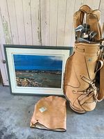 Set of vintage left handed Wilson golf clubs (irons and drivers) in a tan vintage leather golf bag with a leather cover next to a framed lithograph print.