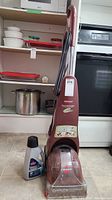 Front view of the Bissell QuickSteamer Powerbrush cleaner with one small bottle of cleaning solution beside it in front of kitchen cabinets and appliances.