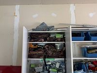 Cabinet with two shelves containing folded camo fabric, hardware boxes, and container of nails; also visible are blue bins with tools beside the cabinet