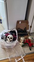 Photo of white storage tote filled with Halloween decorations including skulls and black/red costume pieces, with a box and some string lights on floor