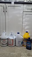 Six bottles of various cleaning solutions arranged on kitchen floor in front of white cabinets.