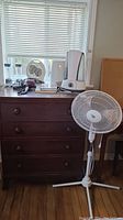 Wide shot showing oscillating pedestal fan, humidifiers, hair dryer, and books on wooden chest.