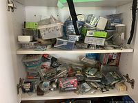 Two shelves inside a white cabinet with assorted fasteners and hardware items stored in plastic containers and packaging, including screws, nails, bolts, and wall plugs.
