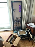 Full length wood framed mirror standing beside multiple stacked picture frames inside a box and on the floor.