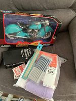Photo of diecast Indian Motorcycle model in turquoise with original box, Cards Against Humanity game box, decks of playing cards, knitting needles, crochet hooks, and packaged yarn on sofa.