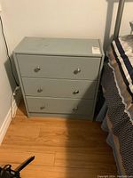 Front view of the small wood nightstand with three drawers alongside a bed. Visible marks on the top surface.