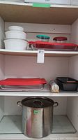 Shelving containing white ceramic casserole dishes with white lids, clear glass Pyrex dishes with red lids, plastic storage containers, marble rolling pin, black bread pans, and a large stainless steel stock pot with lid.