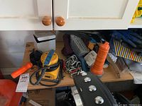 Photo of wooden shelf holding an electric sander with yellow casing, chains, orange wooden clamp, flashlight, and gloves.